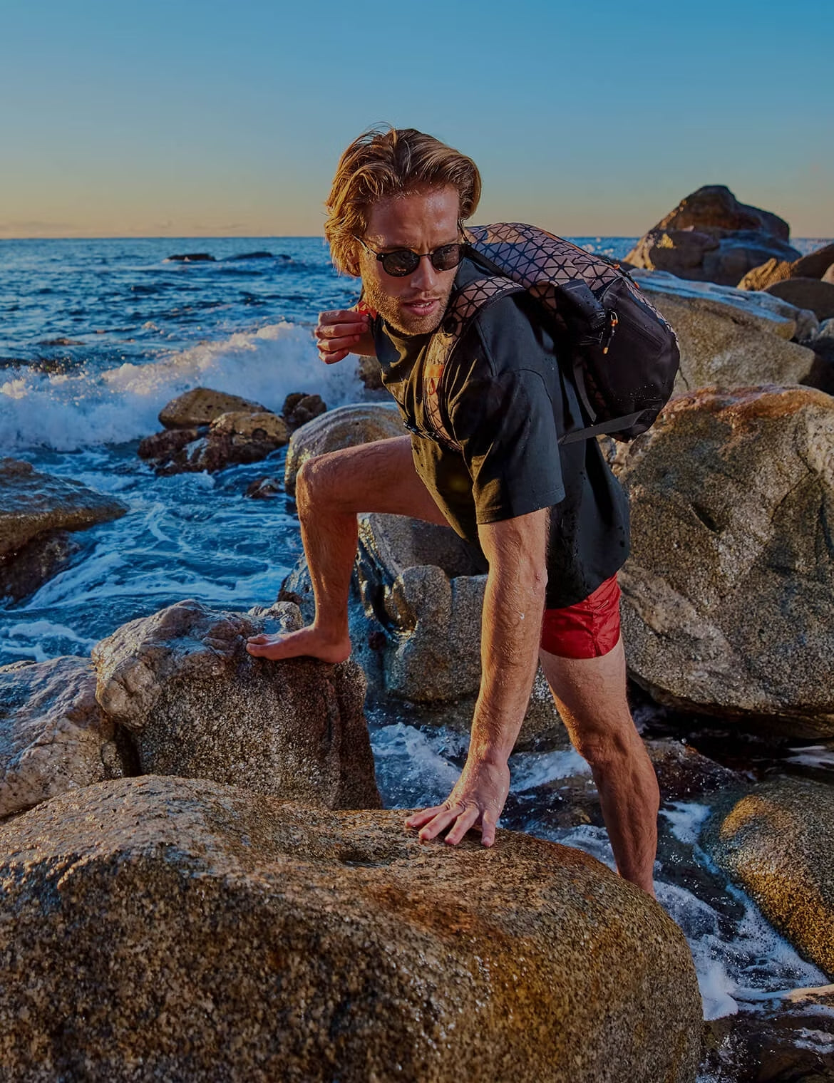Person climbing over wet coastal rocks at sunset, wearing sunglasses, a black shirt, red shorts, and a patterned backpack, with ocean waves behind them.