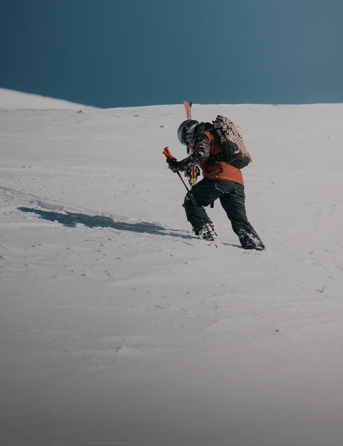 Person climbing a snowy slope using poles, wearing a helmet and carrying skis and a patterned backpack.