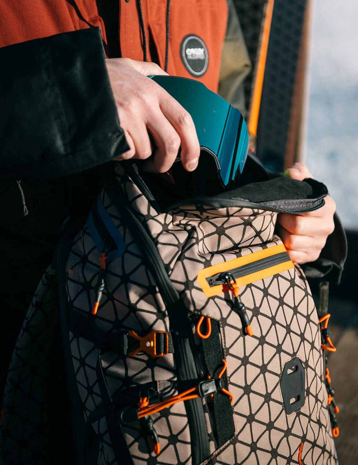 Close-up of hands placing ski goggles into a patterned backpack with orange and black detailing.