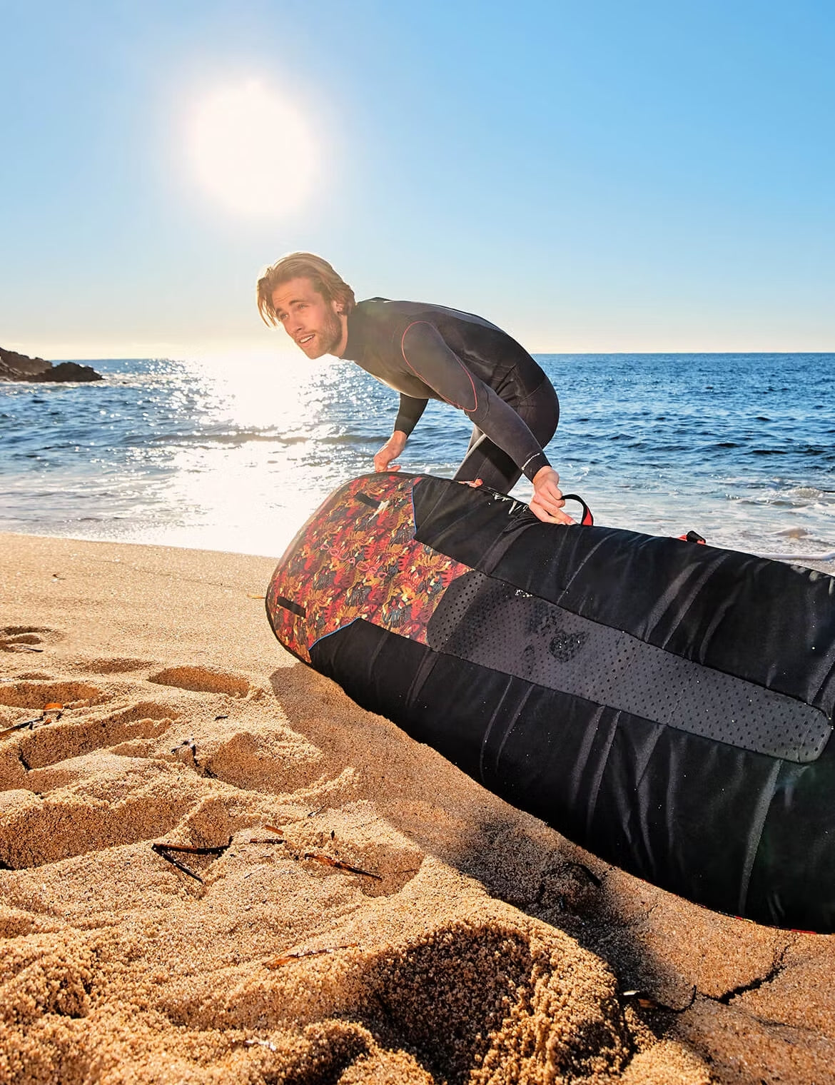 Person in a wetsuit pulling a large padded surfboard bag across sandy beach with bright sunlight reflecting on the ocean behind them.