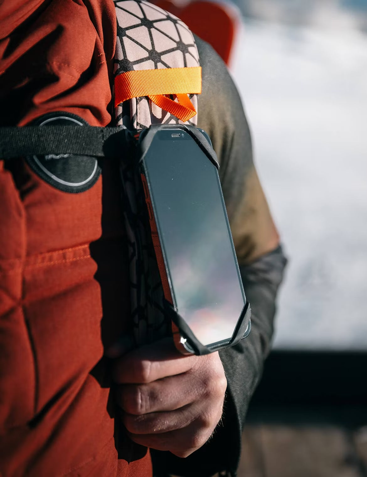 Close-up of a rugged smartphone attached to a backpack shoulder strap with an orange loop, held in place by a person wearing outdoor winter gear.