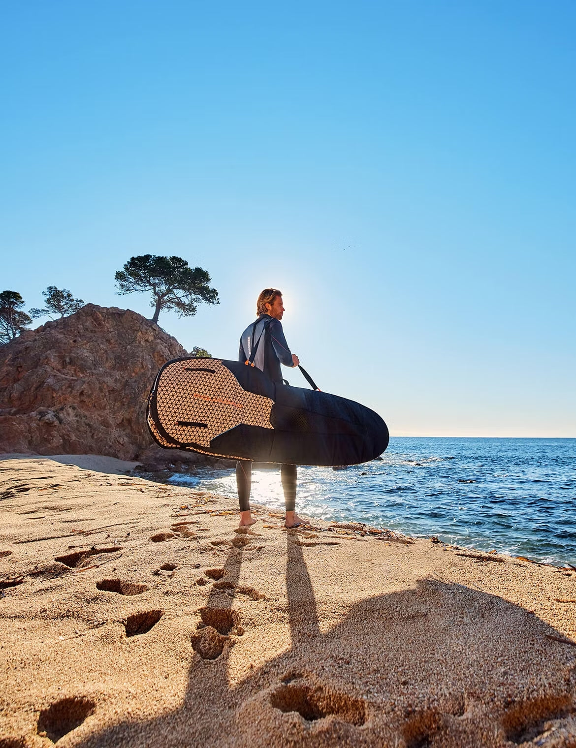 Person in a wetsuit standing on a sandy beach holding a surfboard bag, with long shadows cast toward the camera and the ocean in front of them.