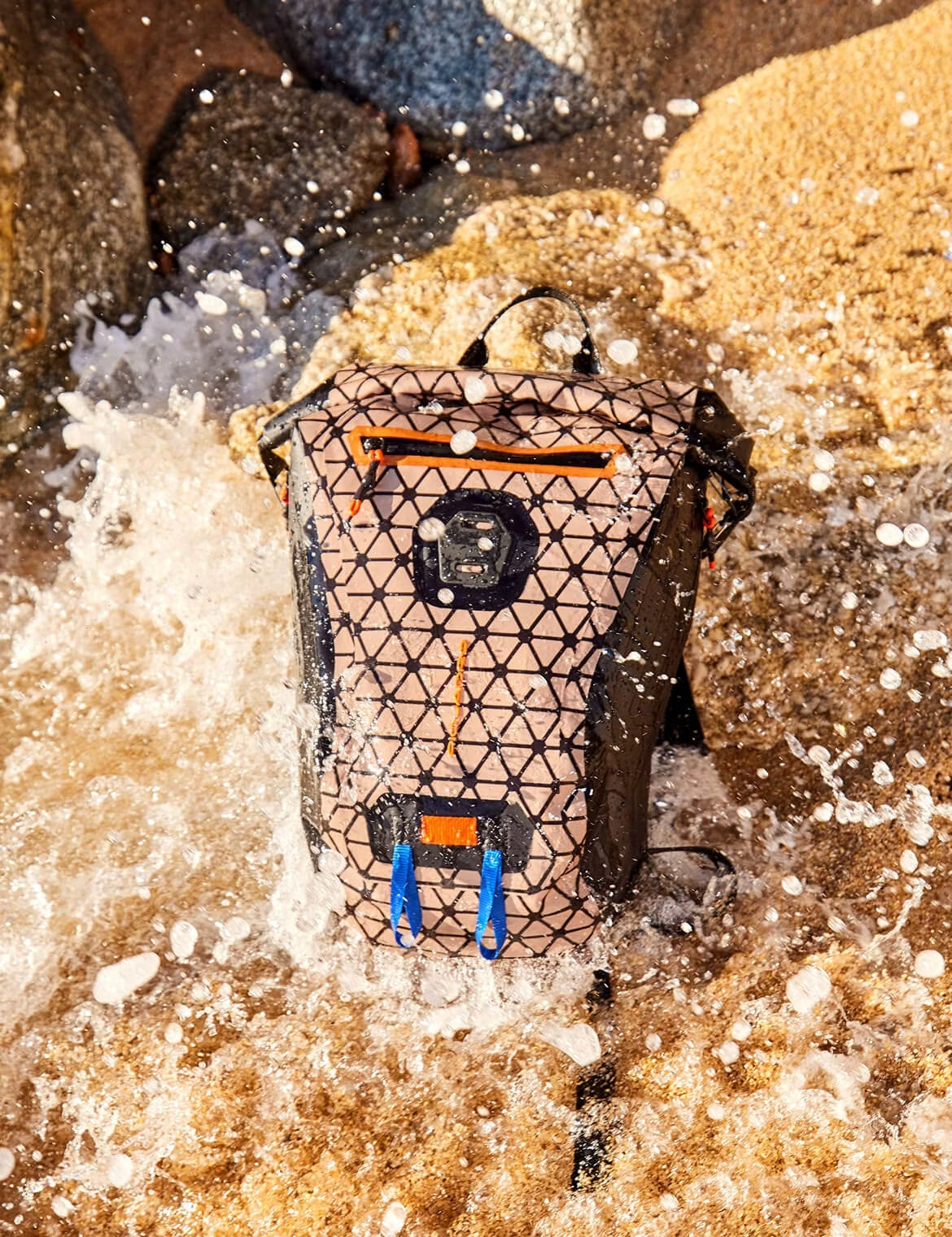Patterned backpack on wet sand being splashed by waves, with water droplets frozen in motion around it.