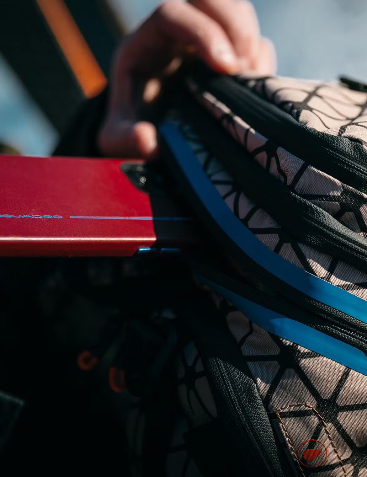 Close-up of a hand placing a red device into the zippered pocket of a patterned backpack with blue and black detailing.