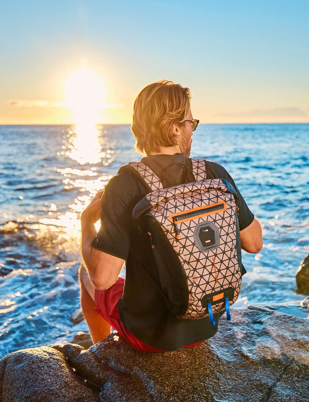Person sitting on coastal rocks at sunset, wearing a patterned backpack and looking out toward the ocean.